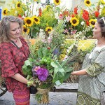 Dwie kobiety pozują do zdjęcia na tle wozu wypełnionego zbożem i kwiatami. Trzymają razem bukiet zielny zawierający fioletowe kwiaty, zioła i zboża.