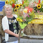 Starsza kobieta trzyma bukiet zielny zawierający słoneczniki, wrotycz, zioła i zboża. Bukiet jest owinięty wstążką w kratkę. Pozuje na tle wozu wypełnionego kwiatami i zbożem.