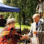 Poseł Bogusław Sonik wręcza torbę z nagrodami laureatce konkursu. Obok nich stoją osoby odpowiedzialne za realizację techniczną wydarzenia. W tle widać figurę świętego Floriana i polanę z drzewami.