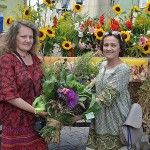 Dwie kobiety razem trzymają bukiet zielny zawierający fioletowe i różowe kwiaty, zioła i zboża. Pozują na tle wozu wypełnionego kwiatami i zbożem.