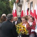 Grupa ludzi ubranych w stroje ludowe wręcza bukiet zielny Prezydentowi Bronisławowi Komorowskiemu i Pierwszej Damie. Za nimi stoi sześć biało-czerwonych flag Polski.