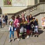 Kobieta i pięć dziewczynek pozują do zdjęcia, trzymają bukiety zielne. Jedna z dziewczynek ubrana jest w strój ludowy. W tle widać Bazylikę Świętego Krzyża w Warszawie i plakat promujący konkurs Cudowna moc bukietów.