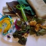 Na stole leżą przygotowane suszone kwiaty i zioła, wstążki, nożyczki i kawałki materiałów.