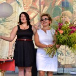 Kobieta z mikrofonem oraz Liliana Sonik stoją na scenie obok stołu z ziołami. Liliana Sonik trzyma duży bukiet zielny. W tle widać obraz przedstawiający procesję zielną.