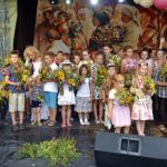 Duża grupa dzieci stoi na scenie, trzymają bukiety zielne. W tle widać obraz przedstawiający procesję zielną.