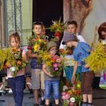 Grupa chłopców i dziewczynka stoją na scenie trzymając bukiety zielne, w tle widać fragment obrazu przedstawiającego procesję zielną.