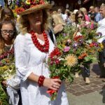 zdjęcie kobiety z koralami na szyi i w kapeluszu, do którego przyczepione zostało dużo kwiatów, trzymającej bukiet zielny. W tle widać dużo osób na Małym Rynku w Krakowie