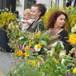 Dwie kobiety, jedna z nich z małym dzieckiem na ręku, trzymają bukiety zielne. Wokół nich widać więcej roślin. W tle znajduje się scena.