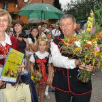 Mężczyzna, kobieta i mała dziewczynka ubrani w stroje ludowe trzymają bukiety zielne. Kobieta trzyma kosz wiklinowy z nagrodą i dyplom. W tle widać zgromadzonych ludzi, jedna z dziewczynek trzyma parasol.