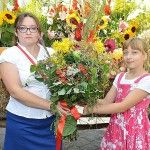 Kobieta i dziewczynka pozują razem do zdjęcia z dużym bukietem zielnym zawierającym jarzębinę, krwawnik i inne zioła i kwiaty. W tle widać wóz wypełniony zbożami i kwiatami.