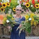 Starsza kobieta pozuje do zdjęcia z dwoma bukietami zielnymi na tle wozu wypełnionego zbożem i kwiatami.