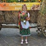 Dziewczynka ubrana w strój ludowy trzyma bukiet zielny, do którego przyczepiono kartkę z numerem konkursowym. Pozuje na tle wozu, do którego przyczepiono baner z napisem "Cudowna moc bukietów, 15 sierpnia 2013 Kraków - Mały Rynek" i logiem Instytutu Dziedzictwa.