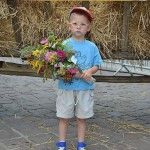 Mały chłopiec pozuje do zdjęcia ze swoim bukietem zielnym na tle wozu wypełnionego zbożem. Do wozu przyczepiono baner z napisem "Cudowna moc bukietów 15 sierpnia 2013, Kraków - Mały Rynek" i logiem Instytutu Dziedzictwa.
