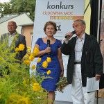 Liliana Sonik i dwóch mężczyzn stoją na scenie. Liliana SOnik i mężczyzna po prawej mówią do mikrofonów. Za nimi stoi pionowy baner z logotypami parterów konkursu. Przed sceną widać roślinę z żółtymi kwiatami.