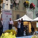 Czterech mężczyzn stoi na scenie na Małym Rynku w Krakowie. Jeden z nich mówi do mikrofonu. Nad ich głowami wiszą kule z kwiatów, a za nimi stoi pionowy baner z logotypami partnerów wydarzenia. Na zdjęciu widać fragmenty bukietów ustawionych pod sceną.