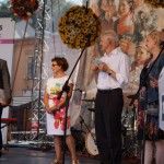 Trzech mężczyzn w garniturze i dwie kobiety stoją na scenie, jedną z nich jest Liliana Sonik. Mężczyzna skrajnie po lewej mówi do mikrofonu, w ręce trzyma teczkę. W tle widać kule z kwiatów powieszone nad sceną.