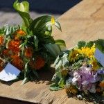 Dwa małe bukiety zielne leżą na stole, mają przypięte karteczki z numerami konkursowymi. Bukiet po lewej składa się z pomarańczowych kwiatów i ziół, a bukiet po prawej z żółtych, różowych i fioletowych kwiatów oraz ziół.
