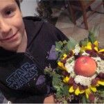 Chłopiec pozuje do zdjęcia z bukietem zielnym stworzonym na warsztatach. W środku bukietu znajduje się jabłko, dookoła niego ułożono kwiaty i zioła.