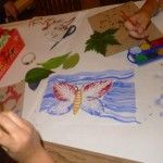 Obrazek motyla z liści na niebieskim w tle. Obok druga osoba maluje swój obrazek przy pomocy farb.