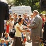 Poseł Bogusław Sonik wręcza laureatce dyplom oraz uściska jej dłoń. W tle widać tłum ludzi zgromadzony podczas wydarzenia, duży głośnik i samochód z napisem "www.wypozyczalniadekoracji.pl".