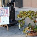 Po lewej stoi tablica z programem konkursu Cudowna moc bukietów w Warszawie. Po lewej stoi stolik, na którym leży dużo kwiatów i ziół. Pod stolikiem znajduje się koszyk wiklinowy wypełniony kwiatami i ziołami.