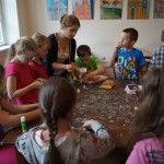 Kobieta pokazuje dzieciom jak zrobić kartki okolicznościowe za pomocą suszonych ziół. Wokół stołu siedzi grupa dzieci.