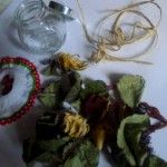Na stole leżą przygotowane suszone kwiaty i zioła, sznurek, materiałowa chusteczka i słoiczek.
