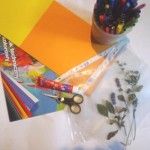 Na stole leżą suszone kwiaty lawendy w koszulce foliowej, nożyczki, klej, mazaki i blok techniczny z kolorowymi kartkami.
