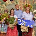 Grupa laureatek i laureat pozują na scenie do zdjęcia z posłem Bogusławem Sonikiem. Kobiety są ubrane w stroje ludowe i trzymają bukiety zielne. Jedna z nich trzyma puchar oraz bilet na wyjazd studyjny do Parlamentu Europejskiego.