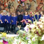Chór Kraków Gospel Choir śpiewa na scenie podczas konkursu. W tle widać obraz przedstawiający procesję zielną. Na pierwszym planie widać nieostre bukiety zielne.