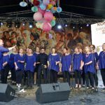Kraków Gospel Choir śpiewa na scenie w czasie konkursu, członkinie ubrane są w granatowe tuniki. W tle widać pionowy baner z logotypami partnerów wydarzenia oraz obraz przedstawiający procesję zielną.