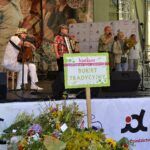 zespół grający na scenie podczas konkursu w Krakowie, w dole zdjęcia widać bukiety zielne złożone na stole oraz tabliczkę "konkurs Cudowna Moc Bukietów Bukiet Tradycyjny"